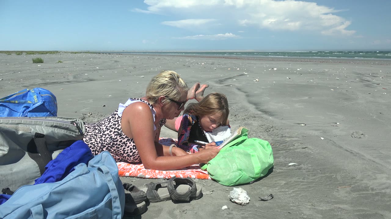 At 0:02, the child keeps their head near the book as the woman lies close on the towel, with a green bag in front and open La Bocana BCS Mexico shoreline behind them.
