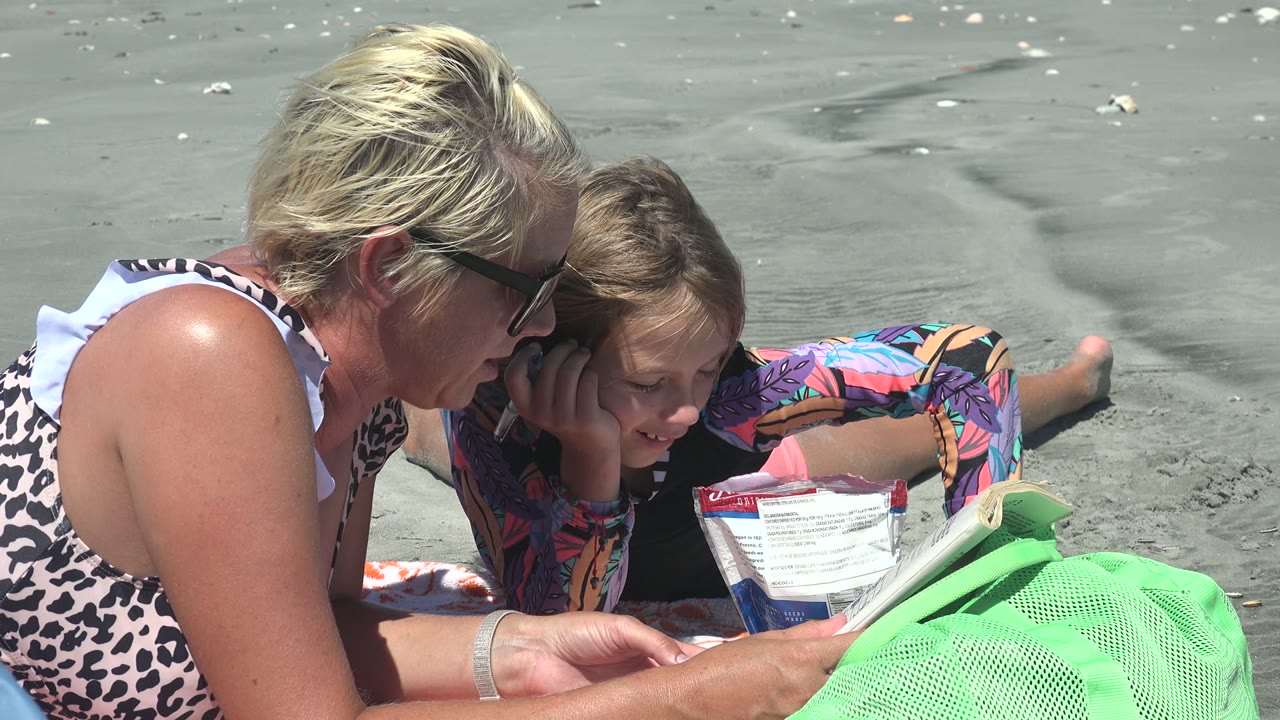 In La Bocana BCS Mexico, a woman in sunglasses lies beside a child on gray sand as both look down toward an object or paper near a green mesh beach bag.