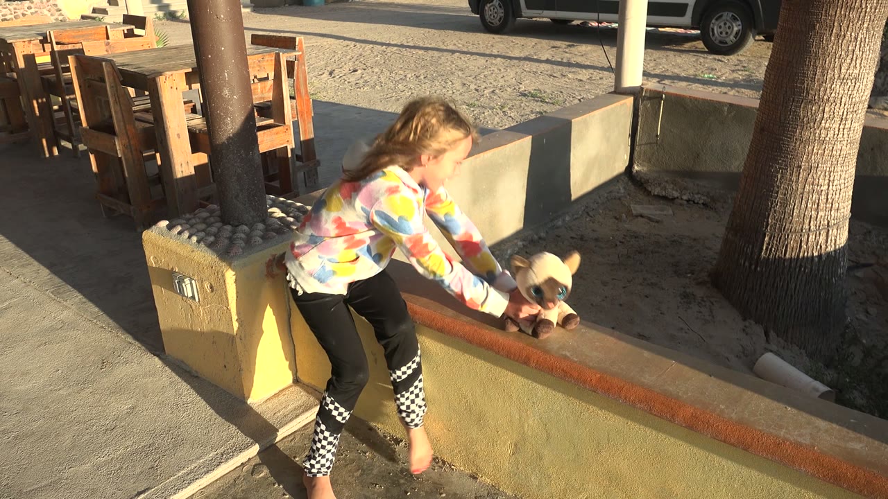 At 0:00 in San Carlos BCS Mexico, a young girl leans over a low yellow patio wall toward a tan stuffed animal, with outdoor tables, chairs, trees, and a parked van in sunlight behind her.