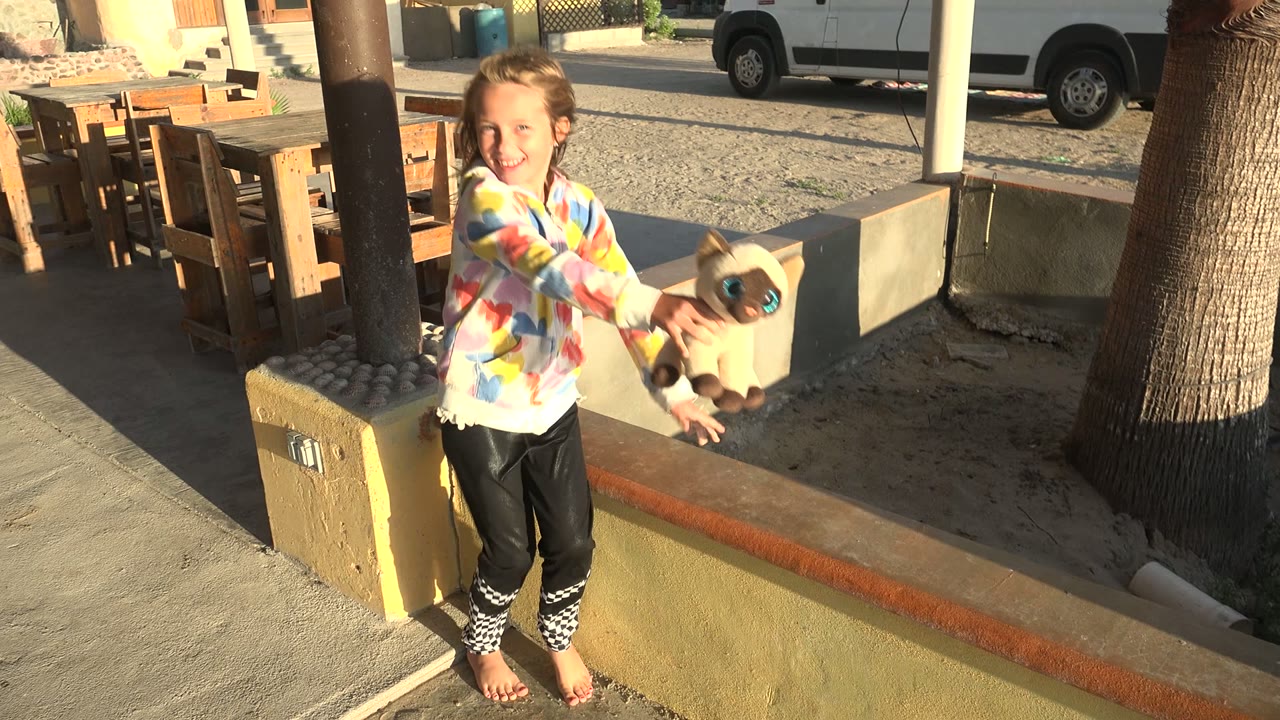 At 0:02 in San Carlos BCS Mexico, the girl stands on the low wall and holds the stuffed animal out in front of her, with bright sun lighting her colorful shirt and the patio furniture behind her.