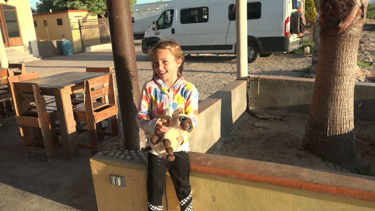 A child in a colorful jacket holds a small light-colored dog or toy-like animal in a San Carlos BCS Mexico courtyard, with wooden furniture and a white van behind them.
