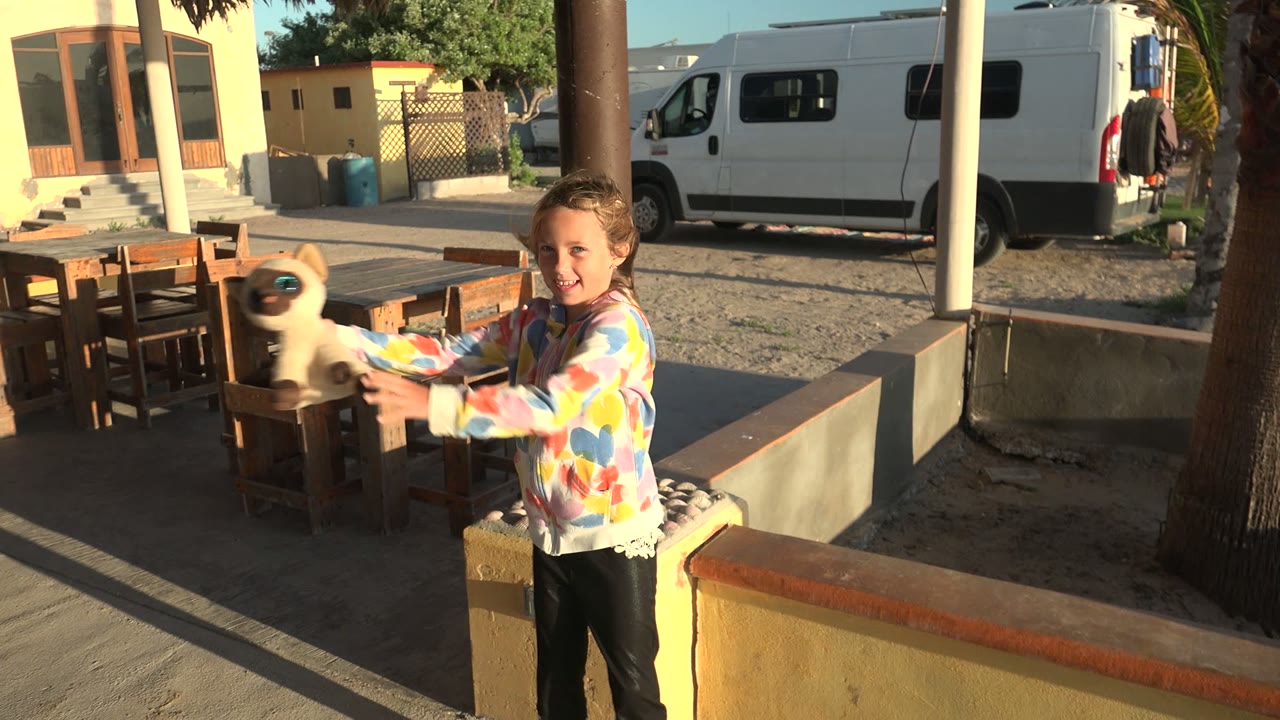 The child extends the small light-colored animal toward the wooden table area in the San Carlos BCS Mexico courtyard, with long shadows crossing the ground.
