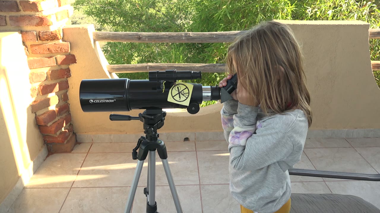 At 0:00, a child in a gray shirt stands beside a black tripod-mounted telescope on a sunlit patio in San Carlos BCS Mexico, with brick columns, a wooden railing, and green foliage behind.
