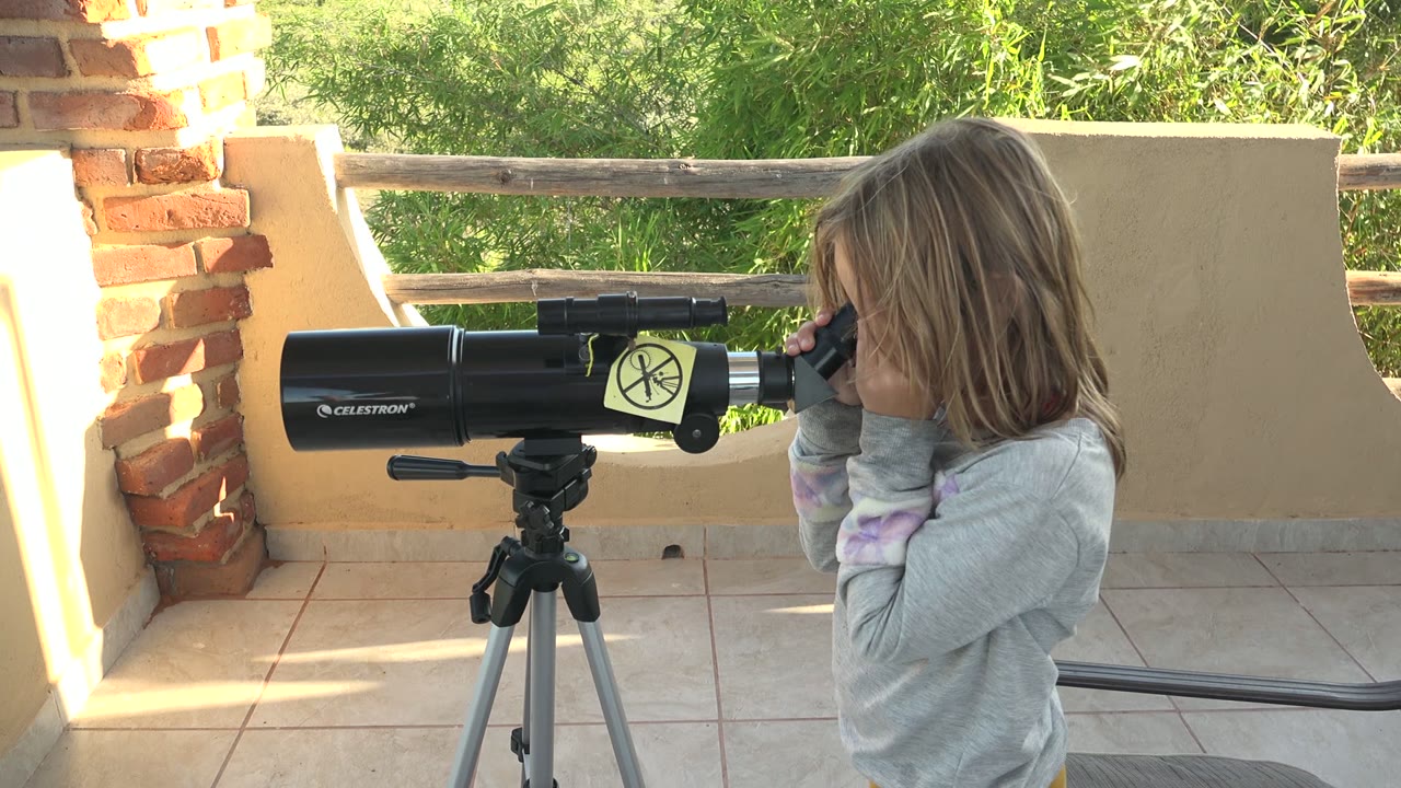 At 0:01, the child remains close to the telescope eyepiece on the tiled patio in San Carlos BCS Mexico, with the black telescope body centered left and the tan wall catching sunlight.