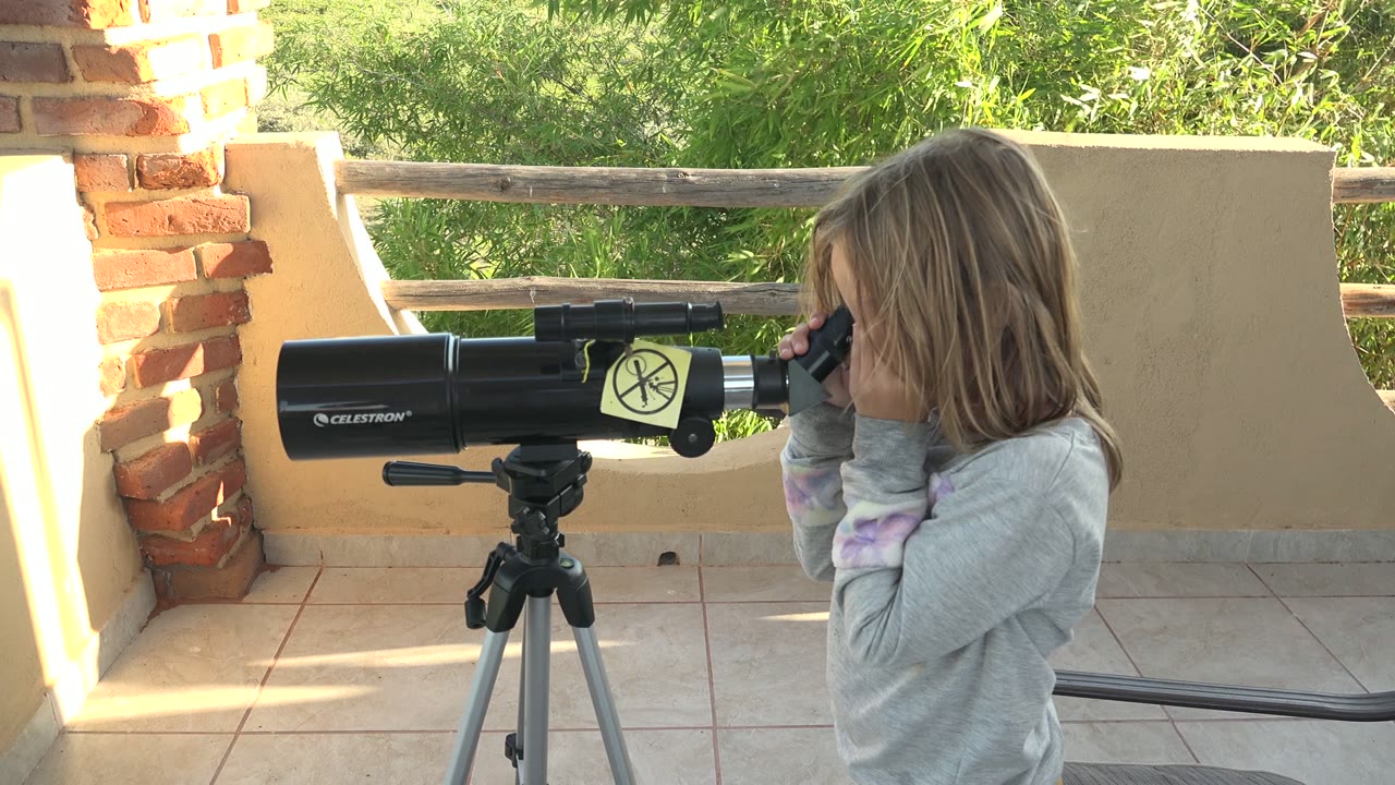 At 0:02, the child continues looking through the black telescope on a tripod in San Carlos BCS Mexico, framed by a brick column at left, a wooden rail, and dense green foliage.