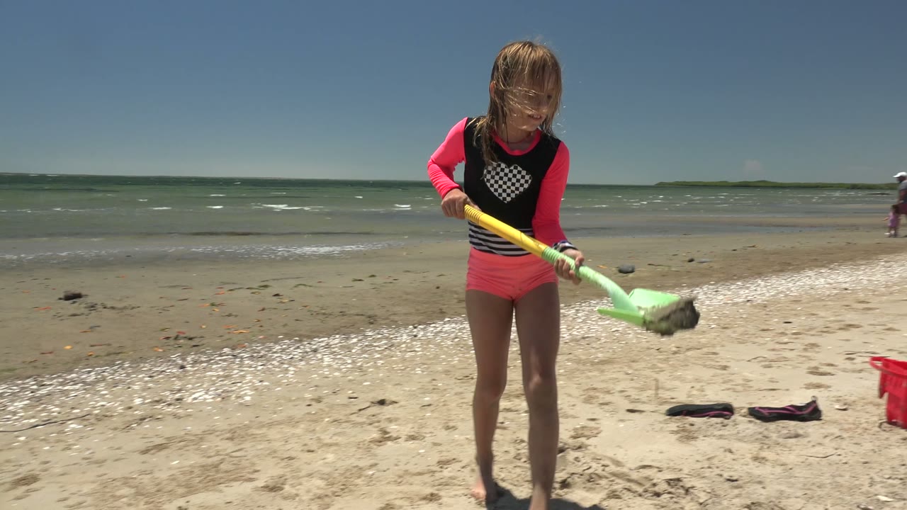 A child in pink and black beachwear digs pale sand with a green shovel on a windy San Carlos BCS Mexico beach, with small waves and distant people behind.