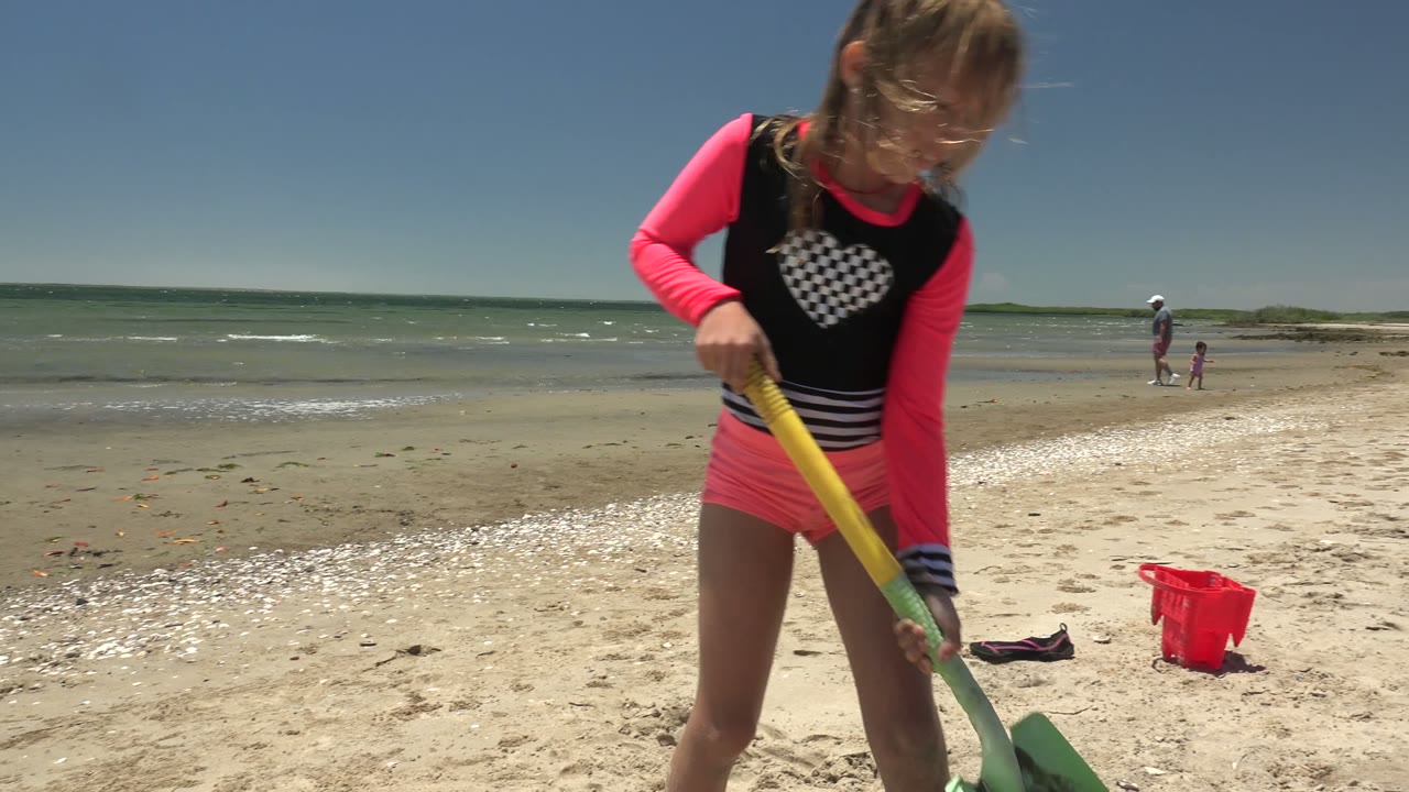 Child leaning over a beach shovel with a red bucket nearby.