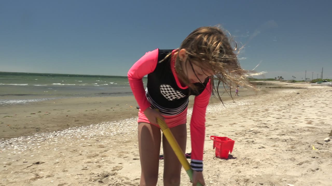 Wind blows the child's hair forward as they bend over the shovel, with the red bucket, scattered beach items, shallow surf, and open sky visible in San Carlos BCS Mexico.