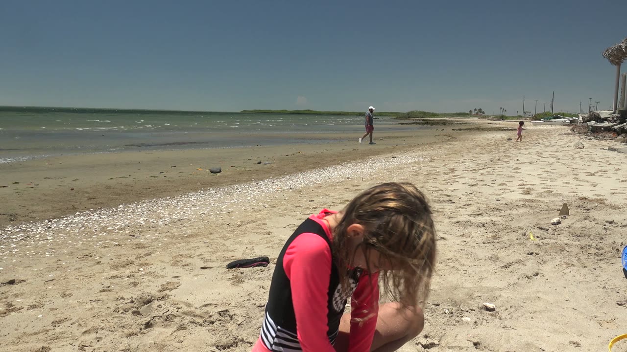 In San Carlos BCS Mexico, the kneeling girl appears on the right foreground with her hair across her face, while the broad sandy shore, ocean, distant beachgoers, boats, and blue sky stay in view.