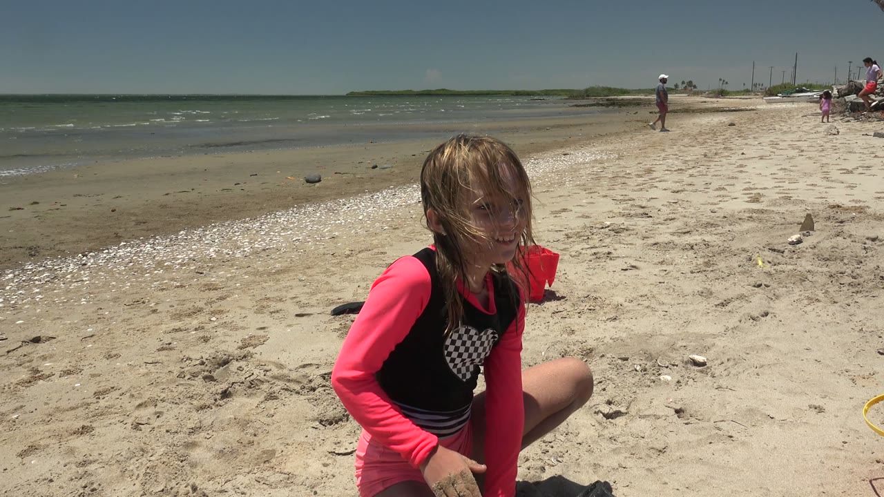In San Carlos BCS Mexico, a girl in a pink and black top crouches in the sand near the lower left while shallow ocean water, shells, and distant beachgoers fill the background.