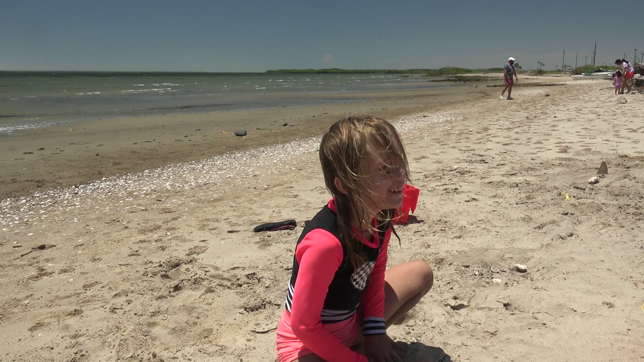 Girl crouched in sand looking downward as waves sit behind her.