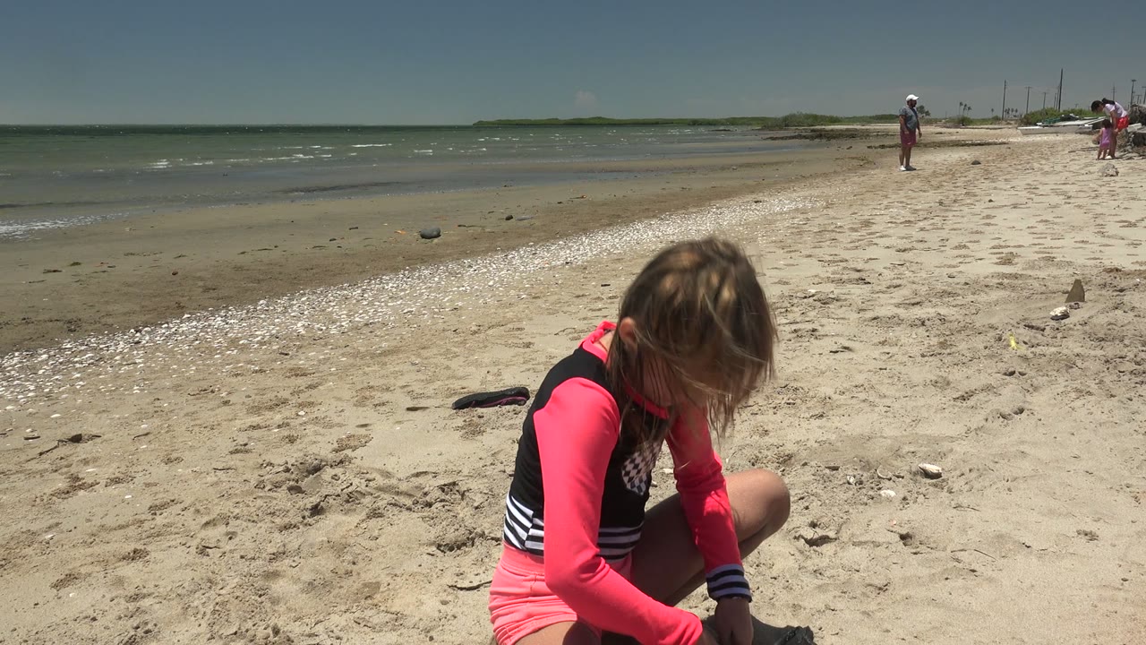 In San Carlos BCS Mexico, the girl leans farther down over the sand in the right foreground while small waves, shells, open beach, and several distant people remain in view.