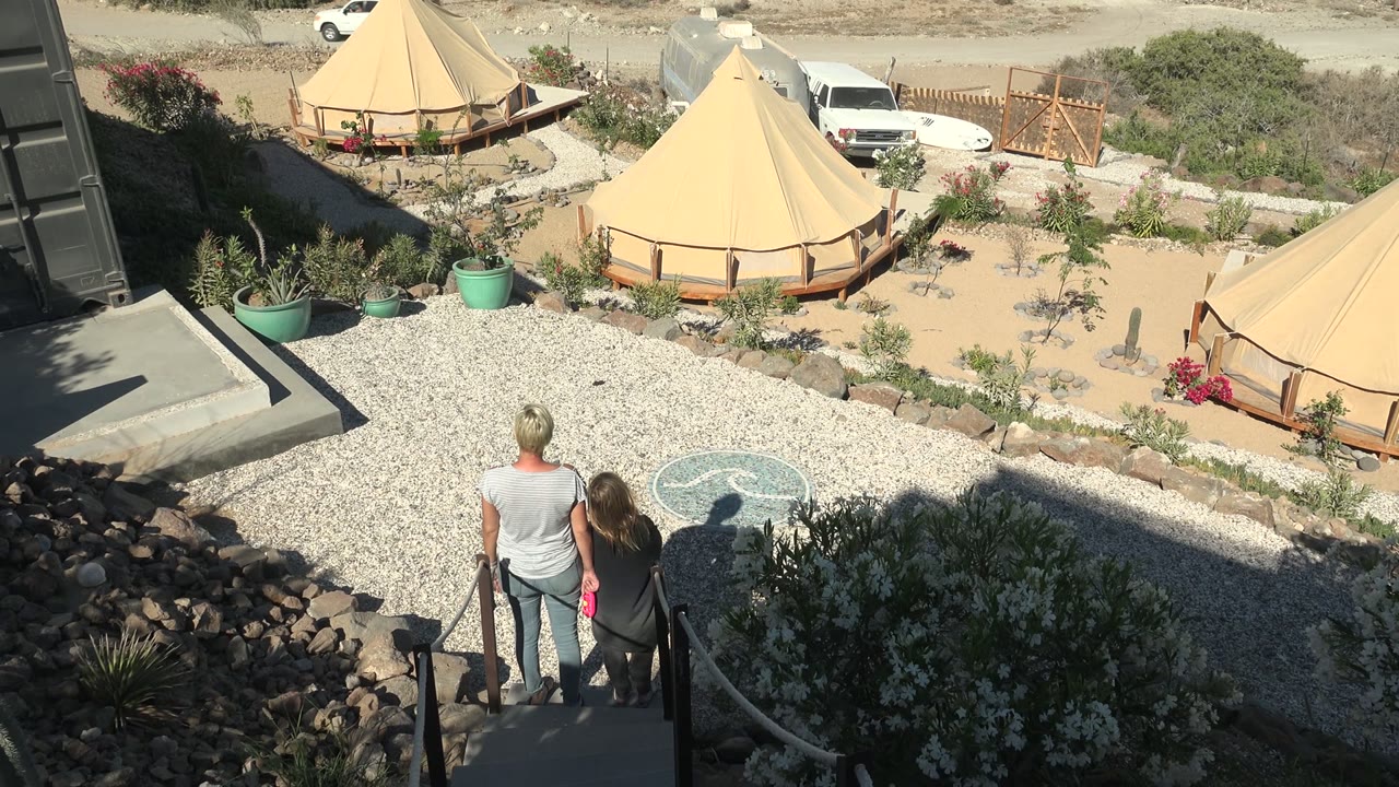 In San Juanico BCS Mexico, a man and woman stand at the steps overlooking a pale gravel courtyard with tan tents, desert plants, rocks, and a white vehicle in the background.