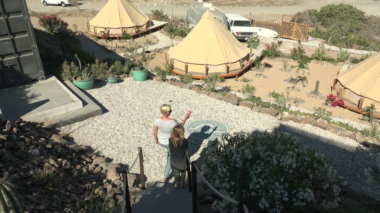 A man in a sleeveless shirt gestures toward the San Juanico BCS Mexico tent area while a woman walks beside him near a circular ground pattern on the gravel.