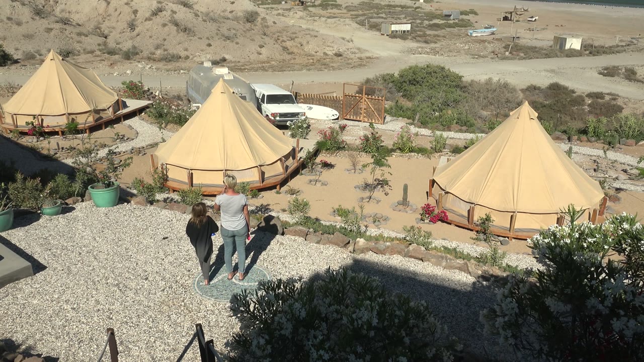 At 0:00 in San Juanico BCS Mexico, two people are visible on a gravel path overlooking tan yurts, desert plants, a sandy beach, and the ocean.