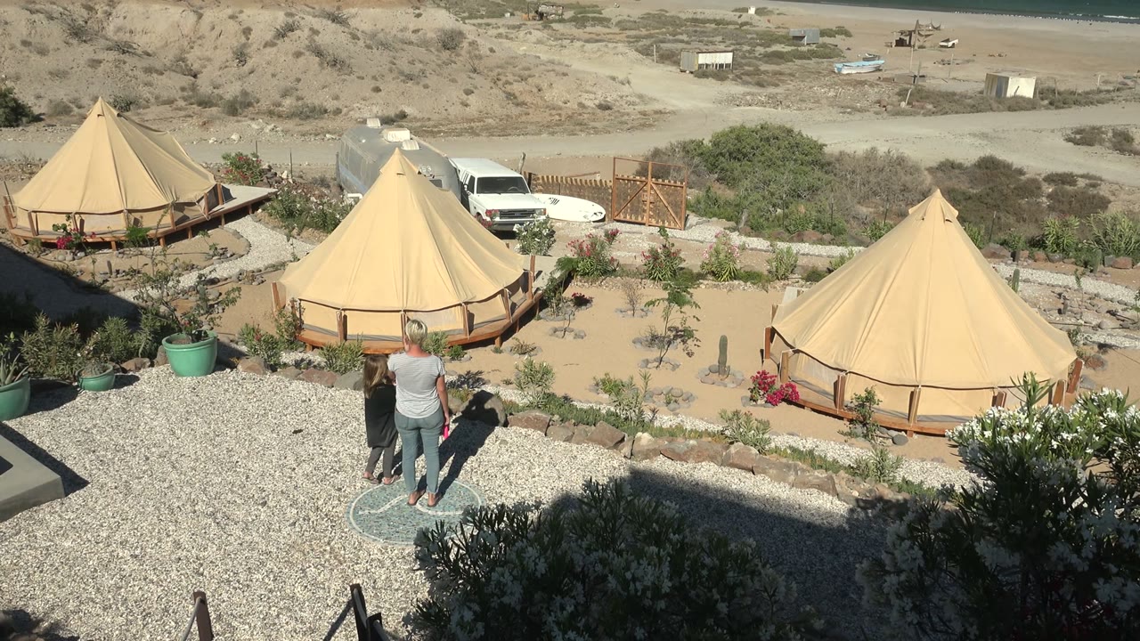 Two people begin walking beside gravel with tan yurts and ocean in the background.