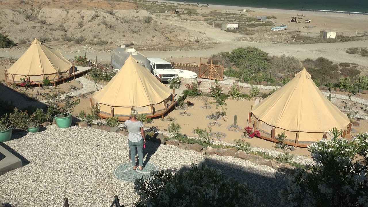 At 0:02 in San Juanico BCS Mexico, one visible person pauses or walks near the gravel edge as the tan yurt roofs, desert planting, parked vehicle, beach, and ocean fill the frame.