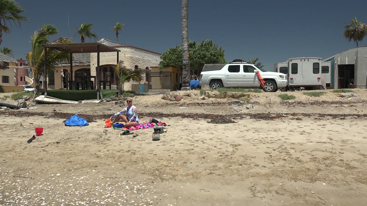 At 0:00 in San Carlos BCS Mexico, a beachgoer sits on a patterned blanket on the sand while a blue bag, red bucket, white truck, homes, and palm trees fill the background.