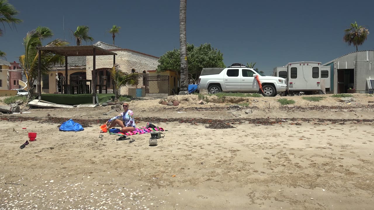 Seated beachgoer adjusts items on sand with a red bucket nearby and a parked white truck behind.