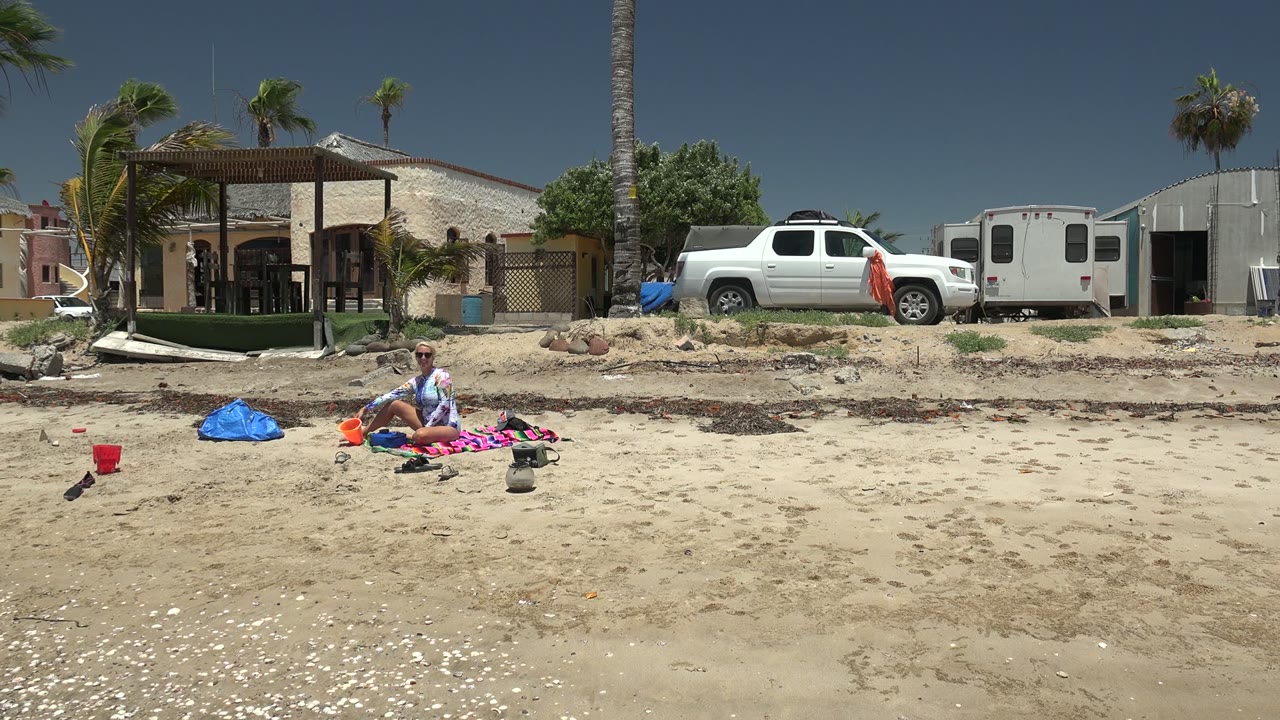 At 0:02 in San Carlos BCS Mexico, the beachgoer sits upright among colorful items on the sand while palms, a white truck, a trailer, and low residential buildings line the rear of frame.