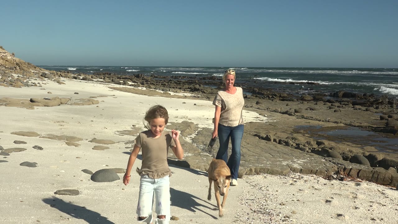 At La Bocana BCS Mexico, a child leads an adult woman and a tan dog across pale sand with rocky shoreline and waves behind them.