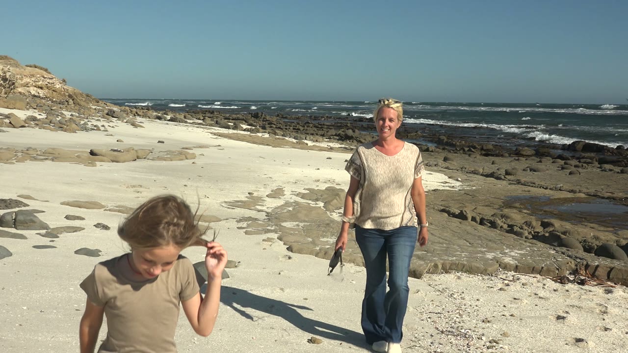 At La Bocana BCS Mexico, the child passes close to the camera as the adult woman follows, with the dog partly low in frame and waves rolling over the rocky shore.