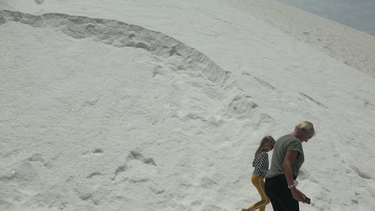 In Guerrero Negro BCS Mexico, a child in yellow pants and an adult in a gray shirt move across a bright white sand dune at the start of the clip.