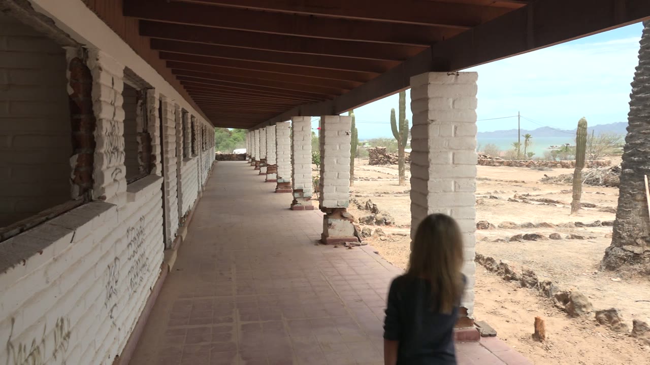 A girl walks away from the camera on a covered walkway in Mulege BCS Mexico, with white brick columns, a tiled floor, sandy ground, cacti, and distant mountains visible.