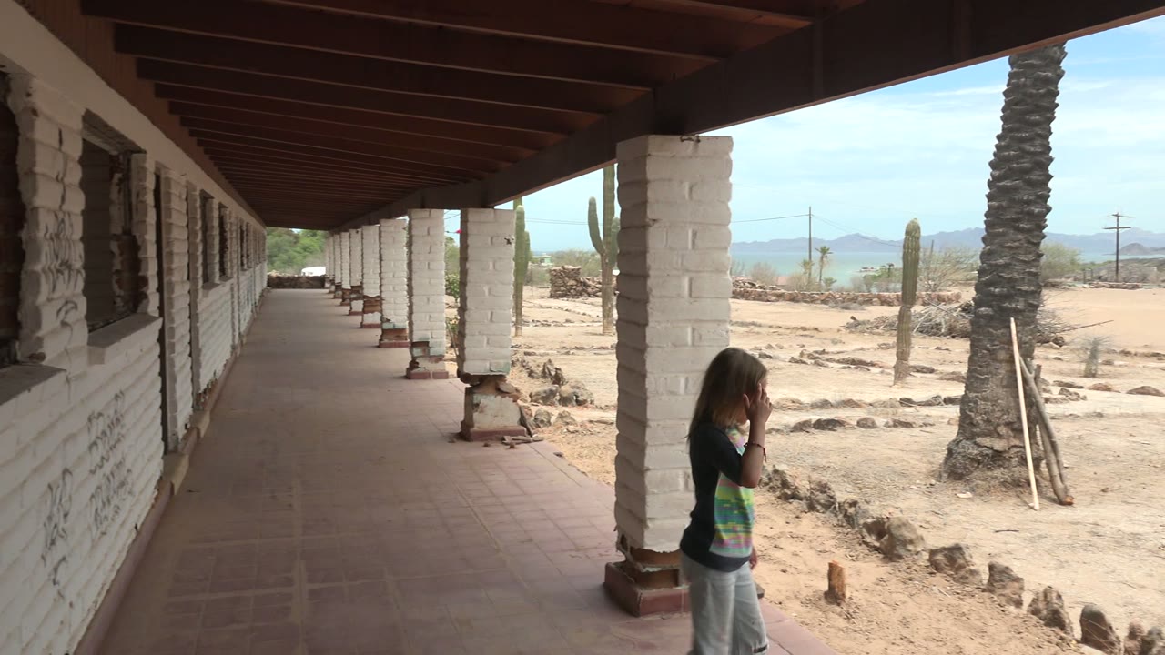 A girl stands near the right edge of the frame on the shaded walkway in Mulege BCS Mexico, with a white column, palm trunk, tall cactus, sandy ground, and low mountains behind her.