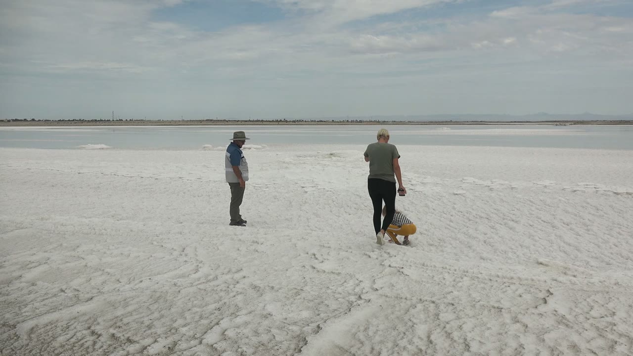 At Guerrero Negro BCS Mexico, two adults stand apart while a child crouches on pale sand near calm water under a partly cloudy sky.