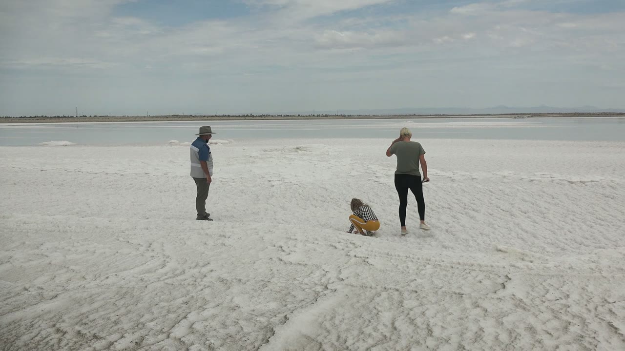 At Guerrero Negro BCS Mexico, one adult stands left of frame as the child crouches and the second adult faces the water on the open sandy shore.