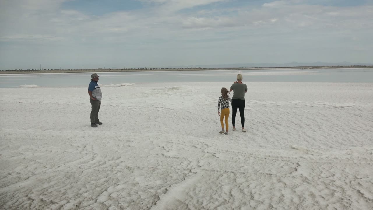Three people are spaced across a white frozen or salt-flat surface in Guerrero Negro BCS Mexico, with shallow water and a distant shoreline under a pale sky.