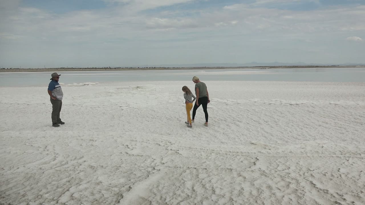 A child and adult move together across the white surface in Guerrero Negro BCS Mexico as another person stands off to the left, with flat water and horizon behind them.