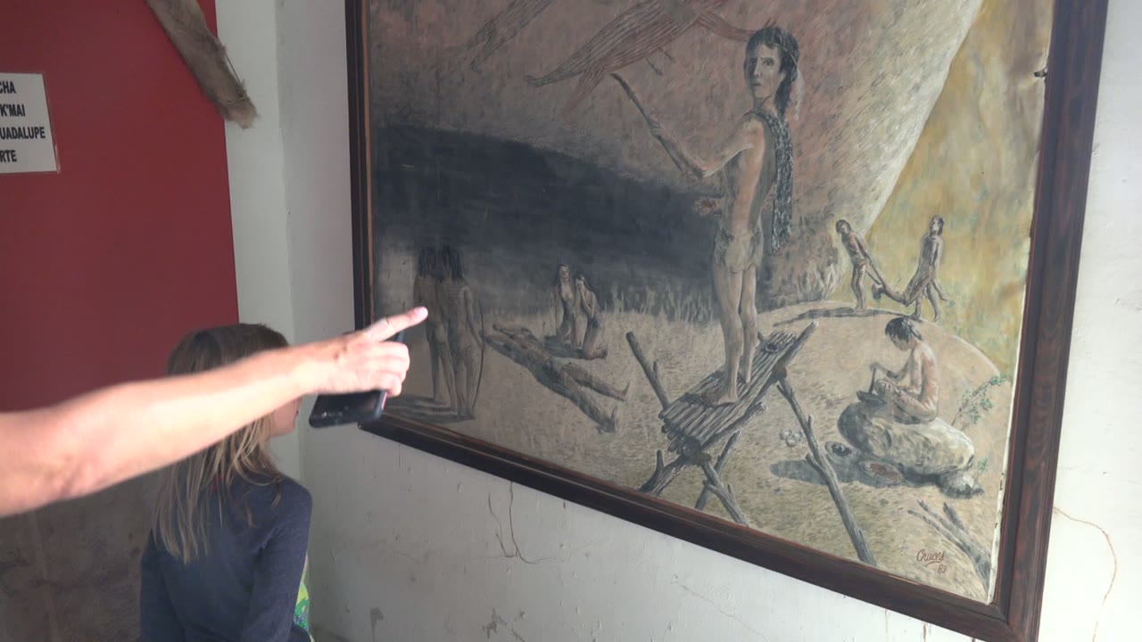 In Mulege BCS Mexico, a girl stands beside a large framed historical scene painting while an adult arm points across the left side of the frame.