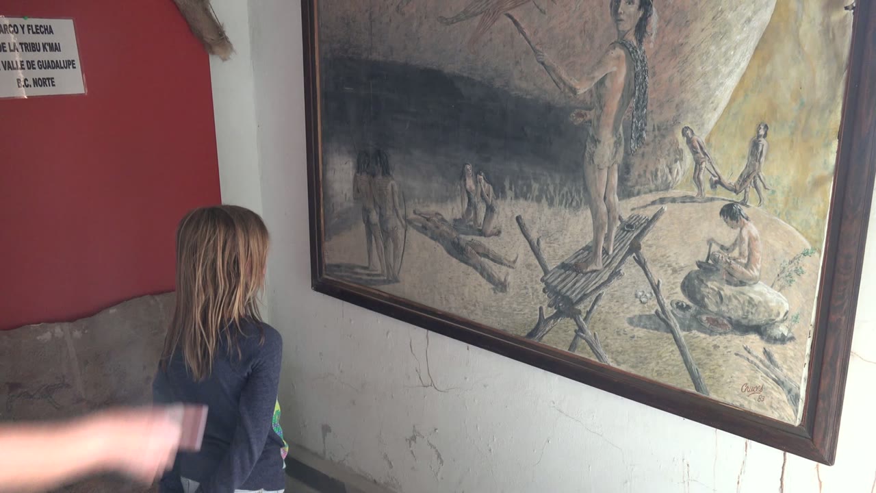 In Mulege BCS Mexico, a girl stands at the left edge of an interior room and looks toward a large framed historical painting on a red and white wall.