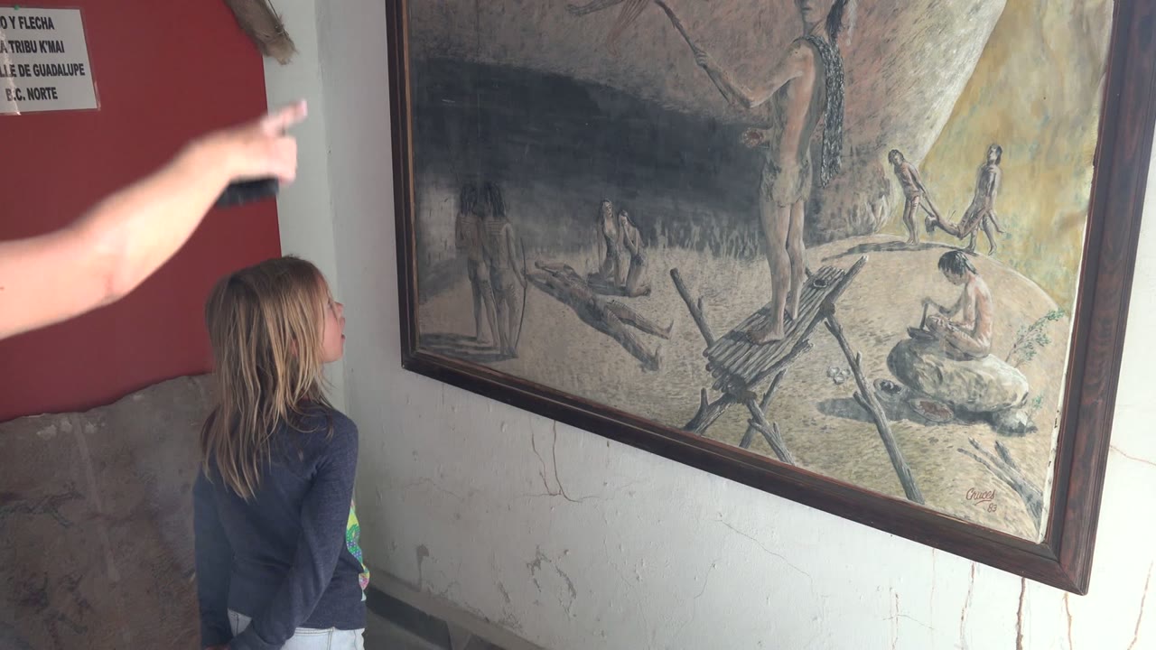 The pointing hand blurs across the upper left as the girl continues watching the large framed historical painting in the Mulege BCS Mexico interior.