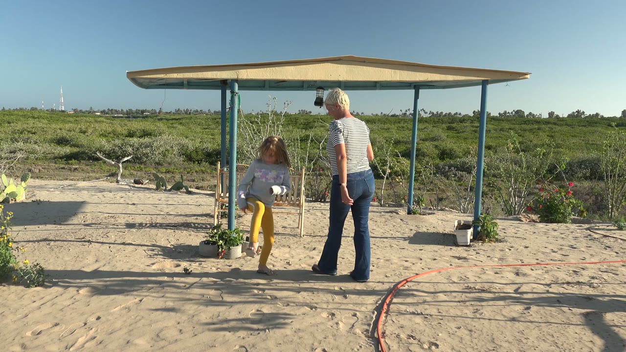 The child stands under the shaded shelter beside the woman in San Carlos BCS Mexico, with potted plants, a small fence, sand, and scrubland visible.
