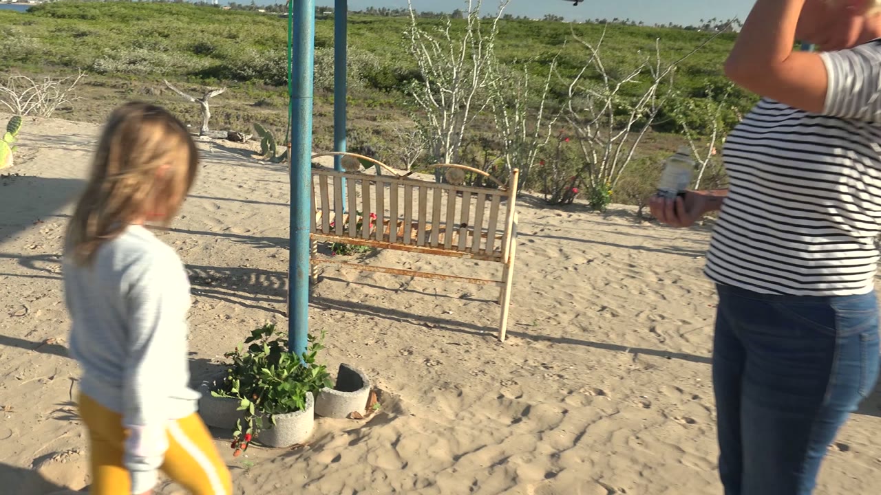 A child in yellow pants walks across sand in San Carlos BCS Mexico near blue poles, a small bench, and a potted plant, with an adult partly visible at the right edge.