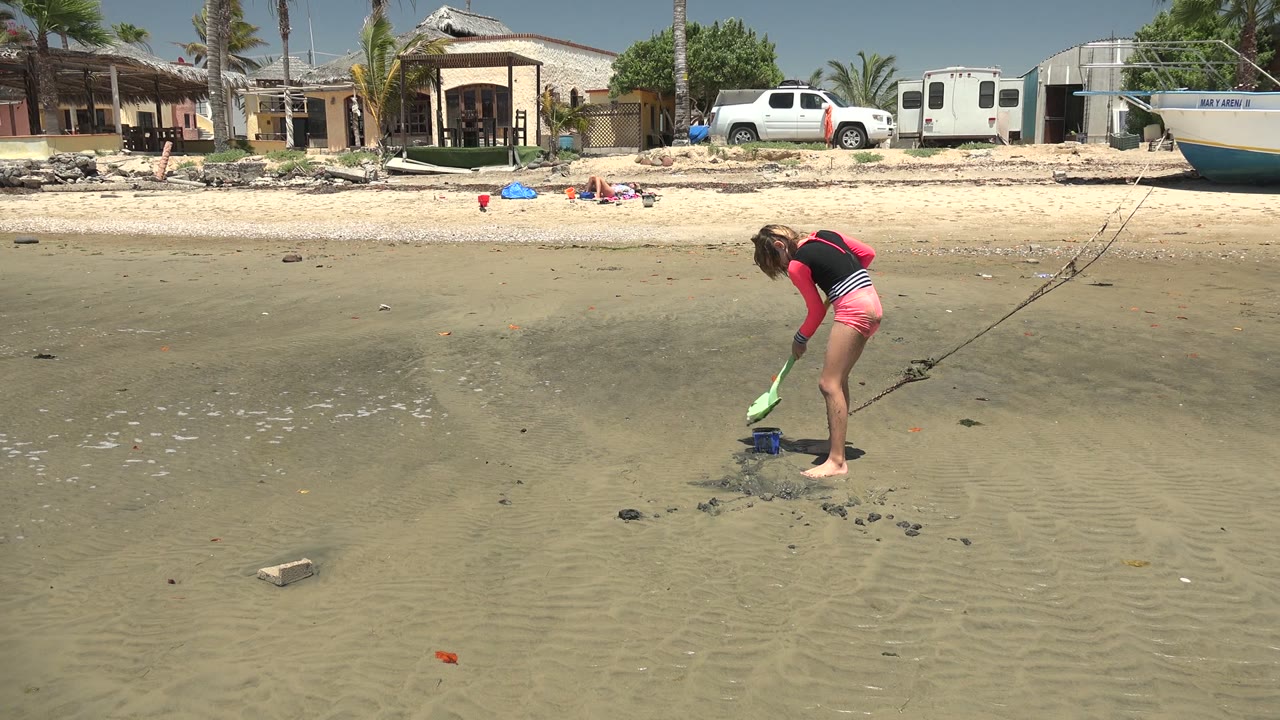 At 0:00 in San Carlos BCS Mexico, a girl in pink shorts bends over wet beach sand with a green shovel while homes, a white truck, a trailer, and a boat sit beyond the shore.