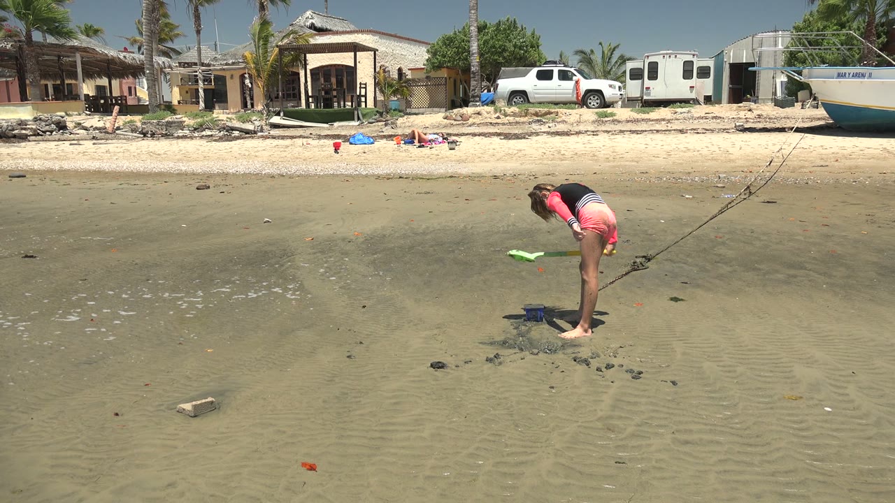 At 0:02 in San Carlos BCS Mexico, the girl shifts the green shovel outward over the wet sand, with shallow water in the foreground and a boat, truck, trailer, and homes in the background.