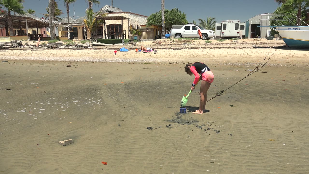 At 0:00 in San Carlos BCS Mexico, a girl bends toward rippled wet sand with a green shovel beside a blue bucket, with a house, white truck, boat, palms, and clear sky in the background.