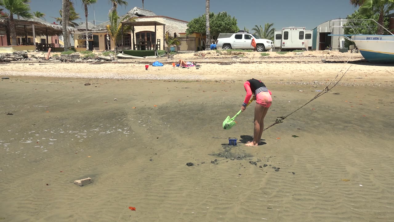 At 0:01 in San Carlos BCS Mexico, the girl holds the green shovel lower toward the wet sand, with the blue bucket nearby and the same beach house, truck, trailer, boat, and palms across the shore.