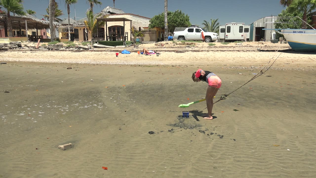 At 0:02 in San Carlos BCS Mexico, the girl leans forward with the green shovel extended away from the blue bucket, while beach homes, a white truck, a small trailer, a boat, palms, and clear sky remain visible.