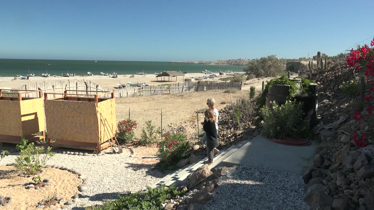 A woman walks along a gravel path in San Juanico BCS Mexico, with desert plants, rocks, a yellow wooden structure, sandy beach, boats, ocean, and clear sky behind her.