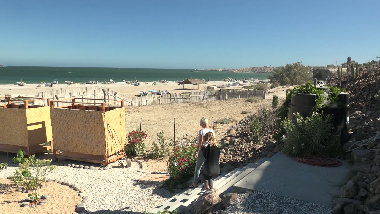 A man joins the woman on the path in San Juanico BCS Mexico, with flowering plants, a dark water tank or planter, rocks, sandy beach, boats, ocean, and blue sky visible.