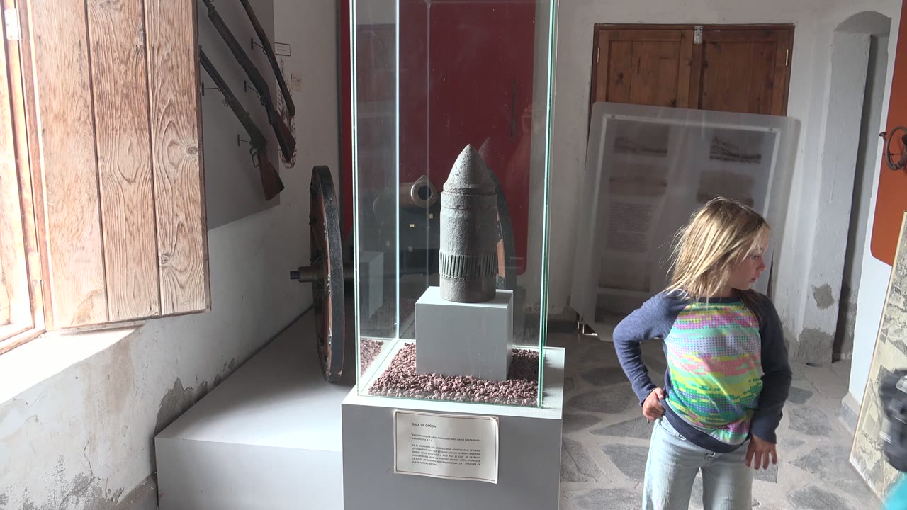 In Mulege BCS Mexico, a young blonde girl stands near a glass case with a dark stone or helmet-shaped artifact, wooden shutters, red wall panels, and stone flooring visible.