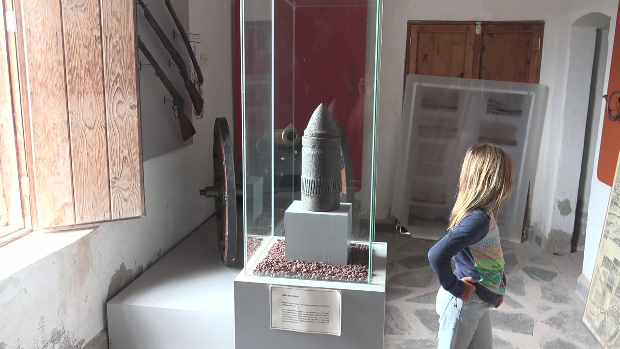 The child is farther to the right of the artifact case, looking away from it while the glass enclosure, dark object, shutters, door, and stone floor remain in view.