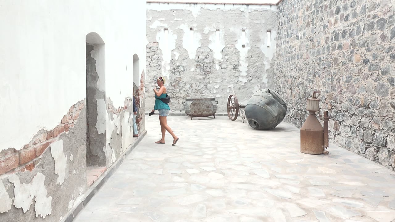 A woman in a blue top and shorts photographs inside a Mulege BCS Mexico stone courtyard, with peeling white plaster, old cannons, wagon wheels, and metal containers visible around her.