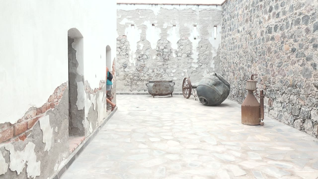 Only part of the woman remains visible at the left doorway in the Mulege BCS Mexico courtyard, with the cannon display, rough stone wall, peeling plaster, and metal container dominating the frame.
