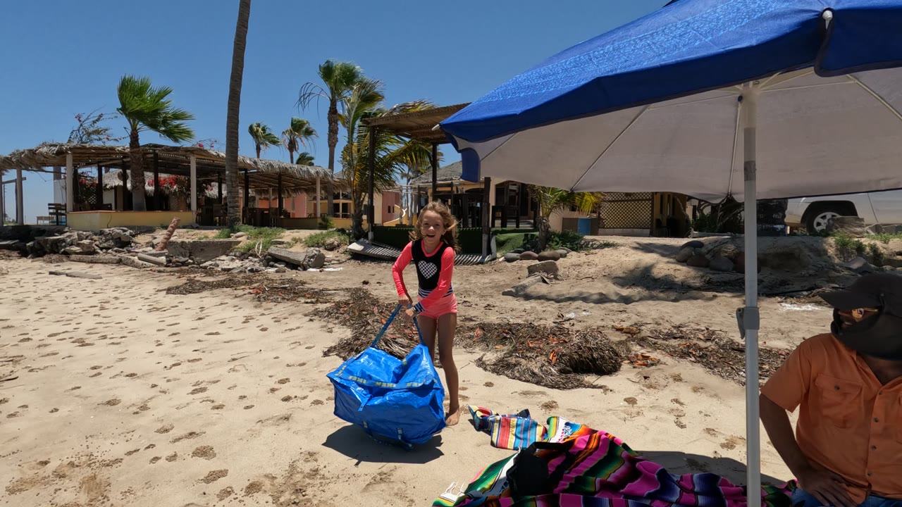 In Baja California Mexico, a child carries a large blue bag across bright sand near a blue beach umbrella, with a striped towel, palm trees, rocks, beach buildings, and a partially visible seated adult nearby.